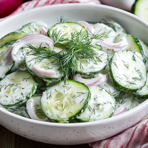 A close-up shot of a bowl of creamy cucumber salad, garnished with fresh dill and thinly sliced red onion, sitting on a wooden table.
