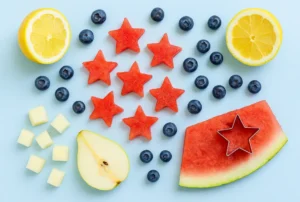 Flat-lay of Patriotic Lemonade ingredients—lemon halves, watermelon stars, blueberries, pear cubes, and a star cutter on a blue background.